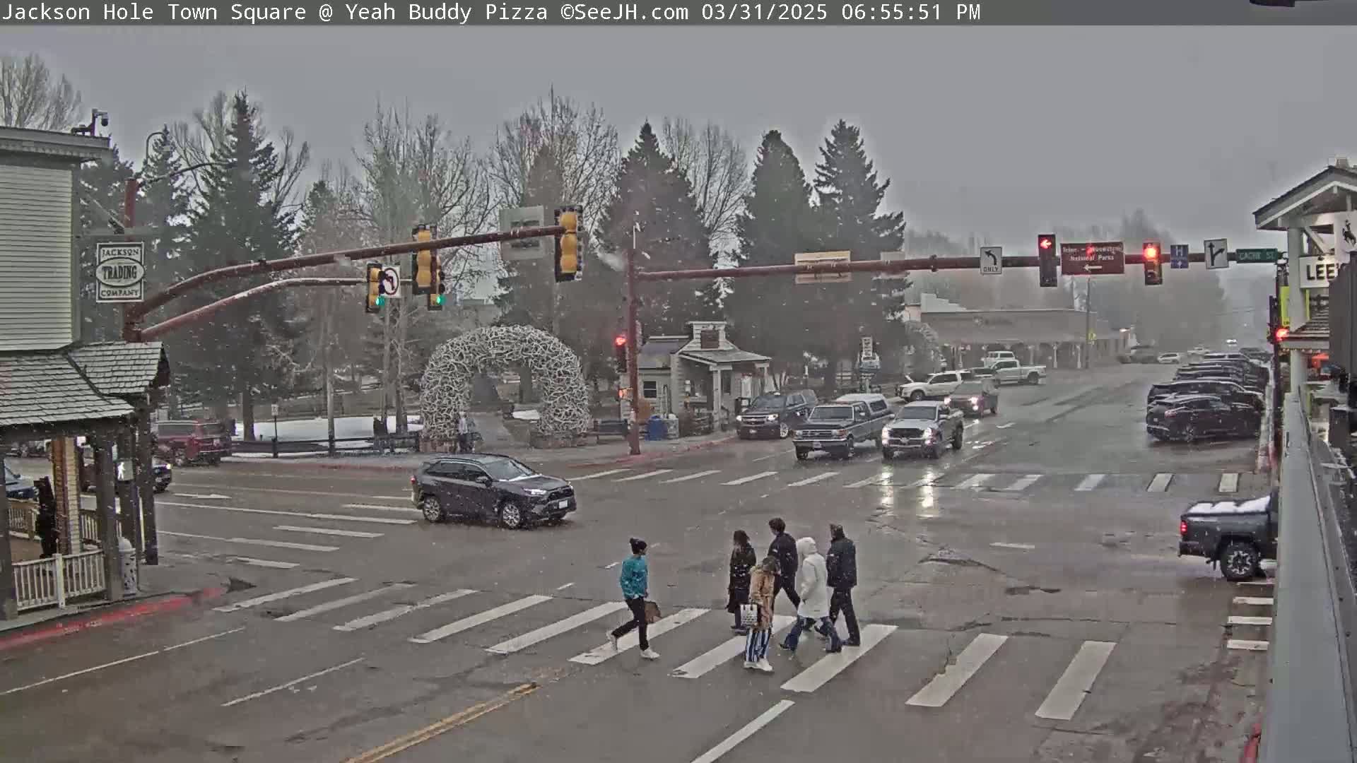 A snowy scene shows a town square intersection with pedestrians crossing in front of cars and buildings, under a gray sky.