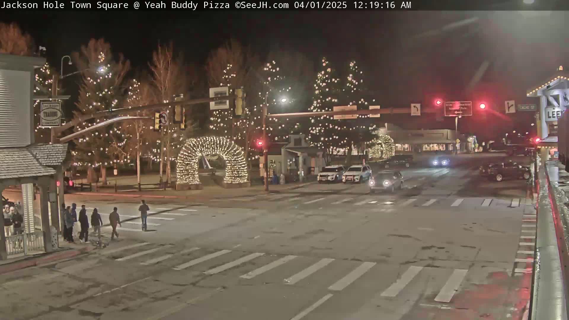 A nighttime street scene shows a town square decorated with Christmas lights, several pedestrians crossing the street, and a few cars at an intersection.