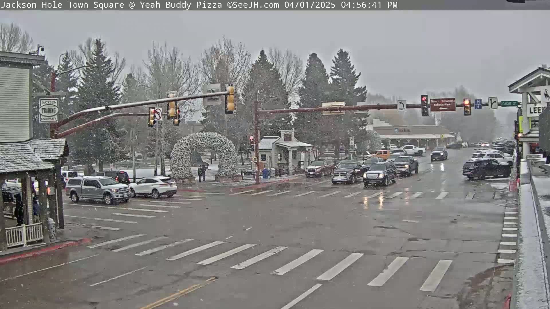A snow-covered town square intersection with several vehicles, traffic lights, and pedestrians, under a light snowfall.