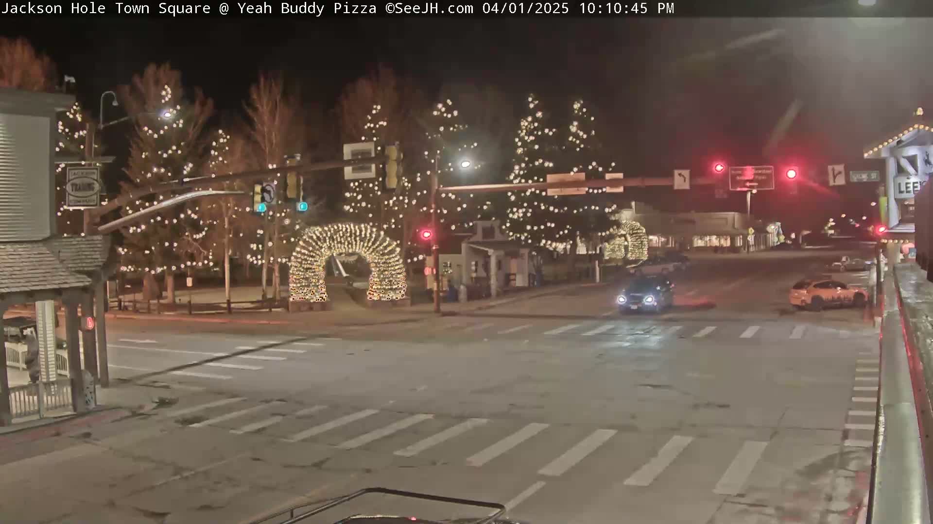 A nighttime street scene shows a town square decorated with string lights on trees and archways, with a few cars at an intersection.