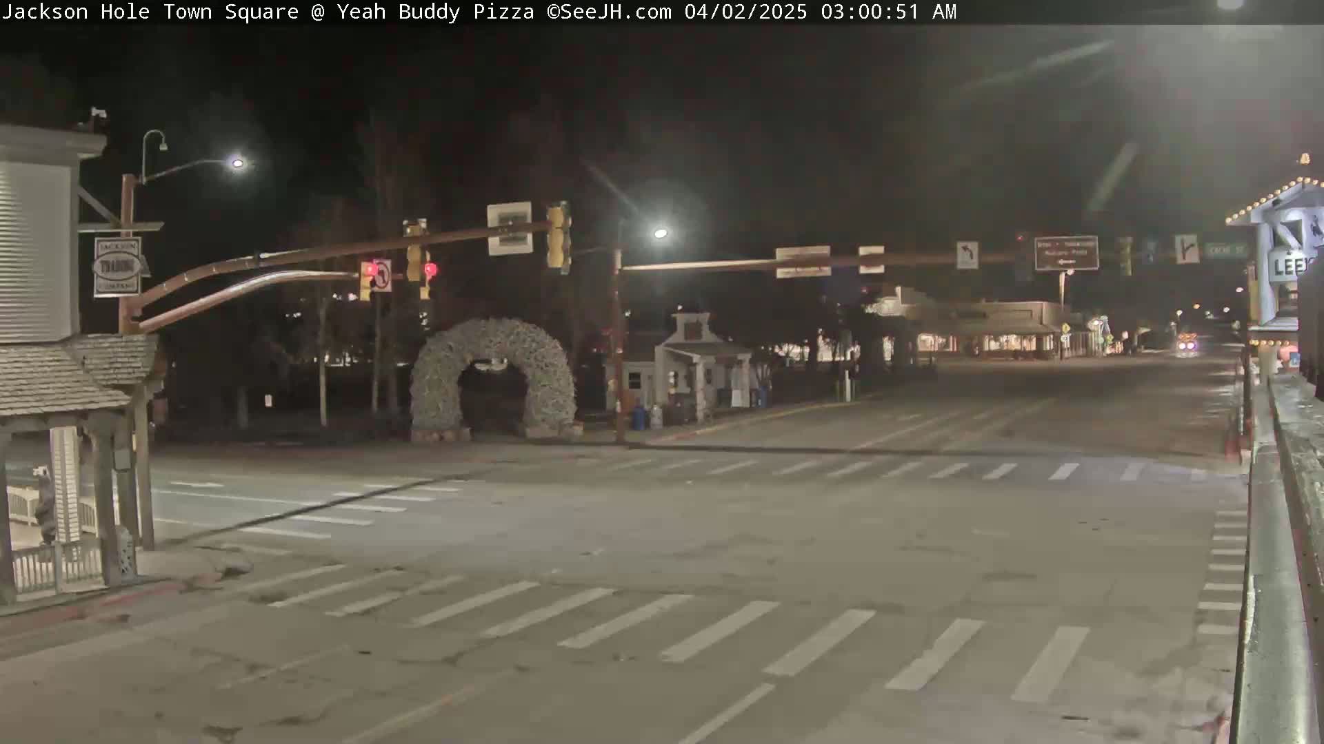 At night, a mostly empty town square intersection shows traffic signals and a large decorative wreath.