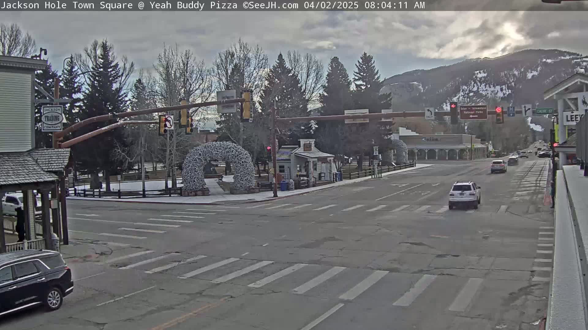 A mostly cloudy day reveals a town square intersection with sparse traffic, snow on the ground, and mountains in the background.
