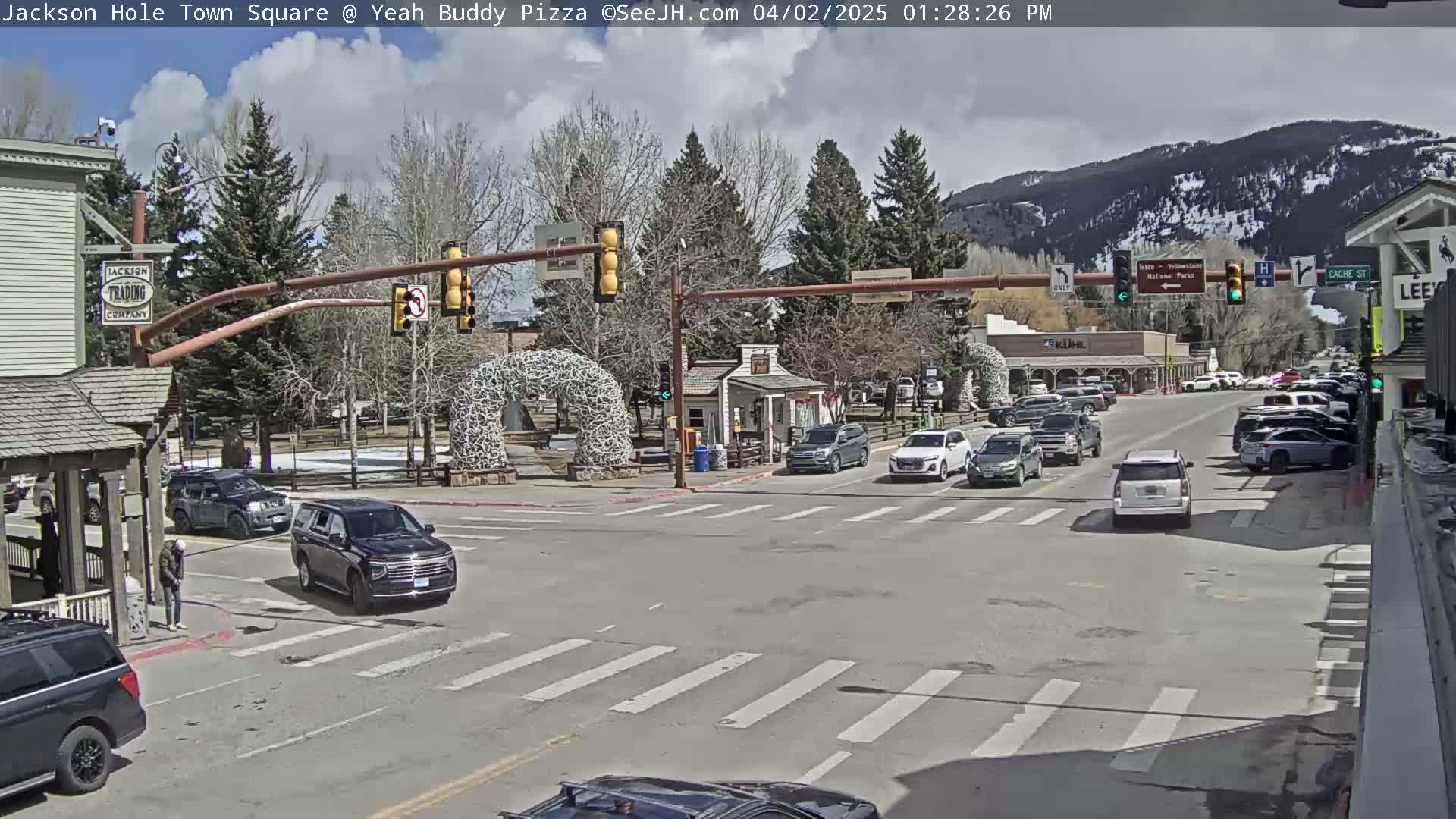 A partly cloudy day in a town square with cars at an intersection, buildings, trees, and mountains in the background.