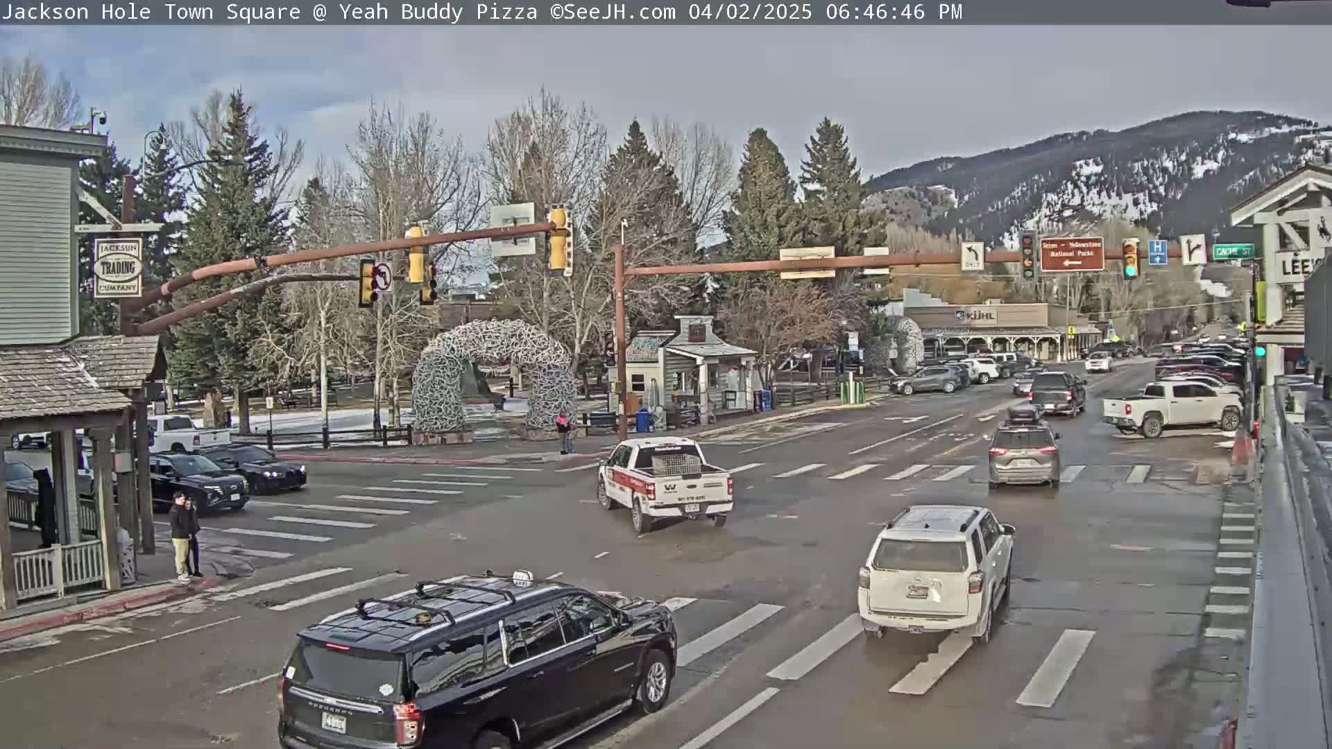 A partly cloudy day shows a town square intersection with several cars and pedestrians, mountains in the background.