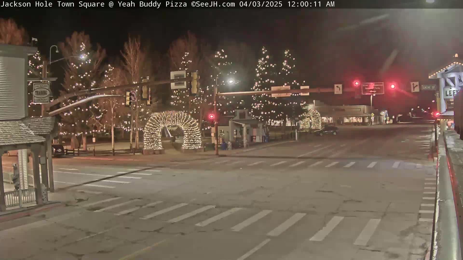 A nighttime street scene shows a town square with trees decorated with lights, an illuminated archway, and traffic signals.