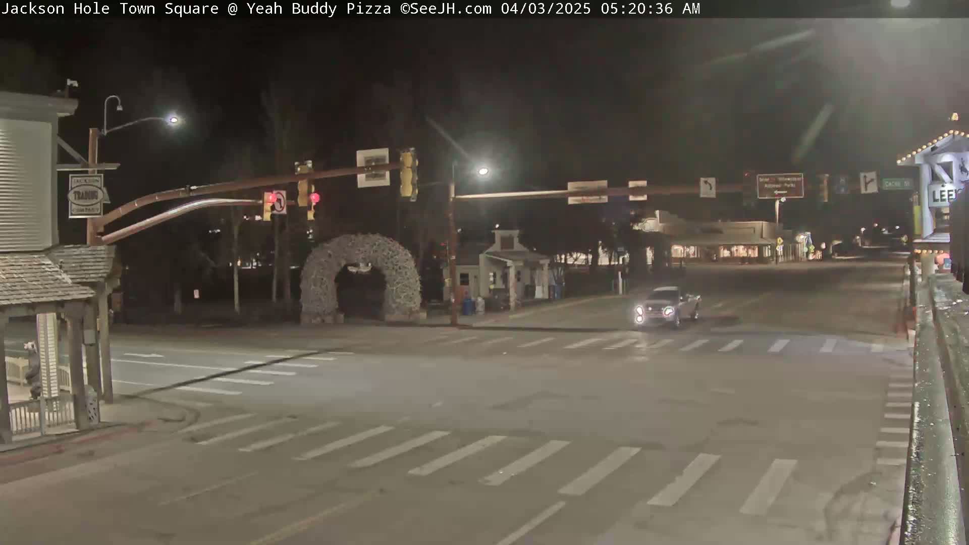 A nighttime view of a town square intersection with a single car crossing, a decorative archway, and buildings lining the street.