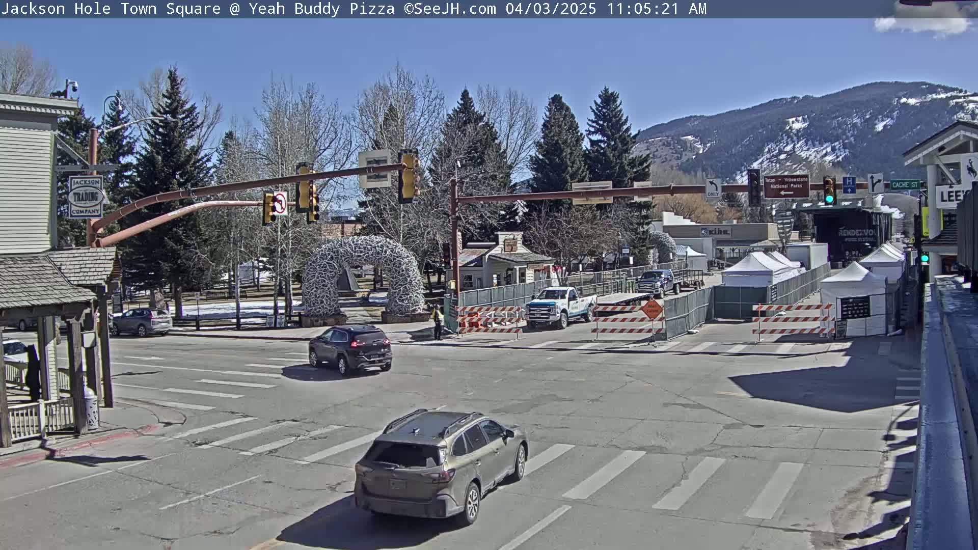 A sunny day reveals a town square intersection with several cars, temporary construction barriers, and white tents, situated near buildings and trees against a backdrop of snow-capped mountains.