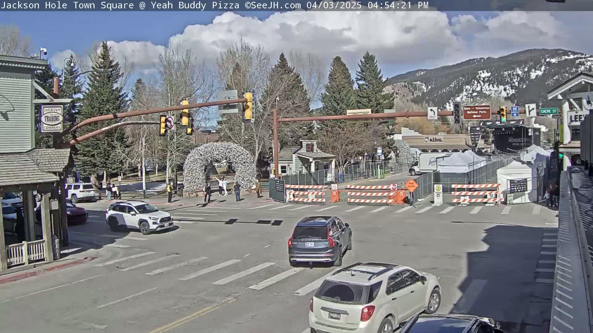 A partly cloudy day shows a town square with several vehicles at an intersection, partially blocked by construction barriers and temporary structures, near a large antler arch.