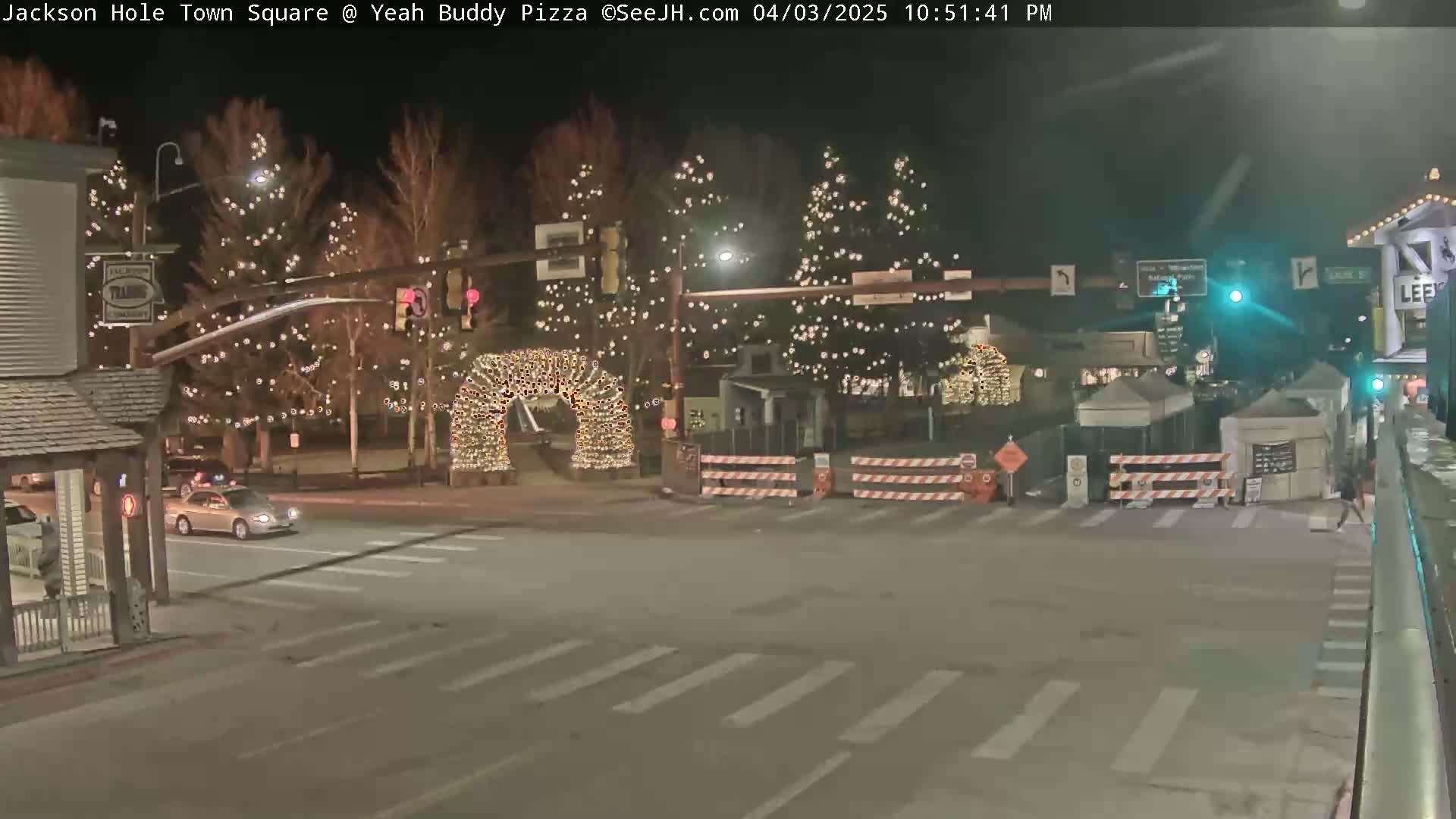 A nighttime street scene shows a town square decorated with Christmas lights, temporary barriers, and a few vehicles.