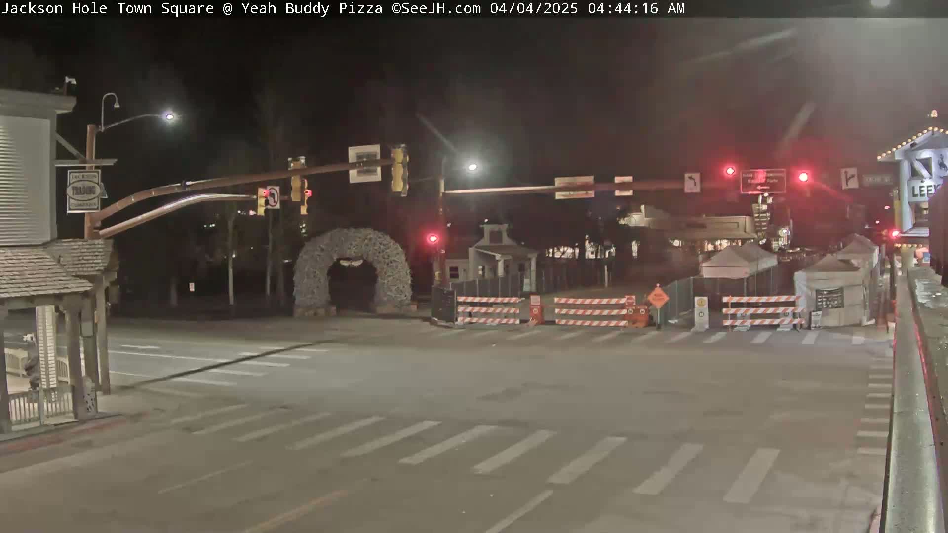 A nighttime view of a town square intersection with traffic signals, construction barriers, and a decorative archway.
