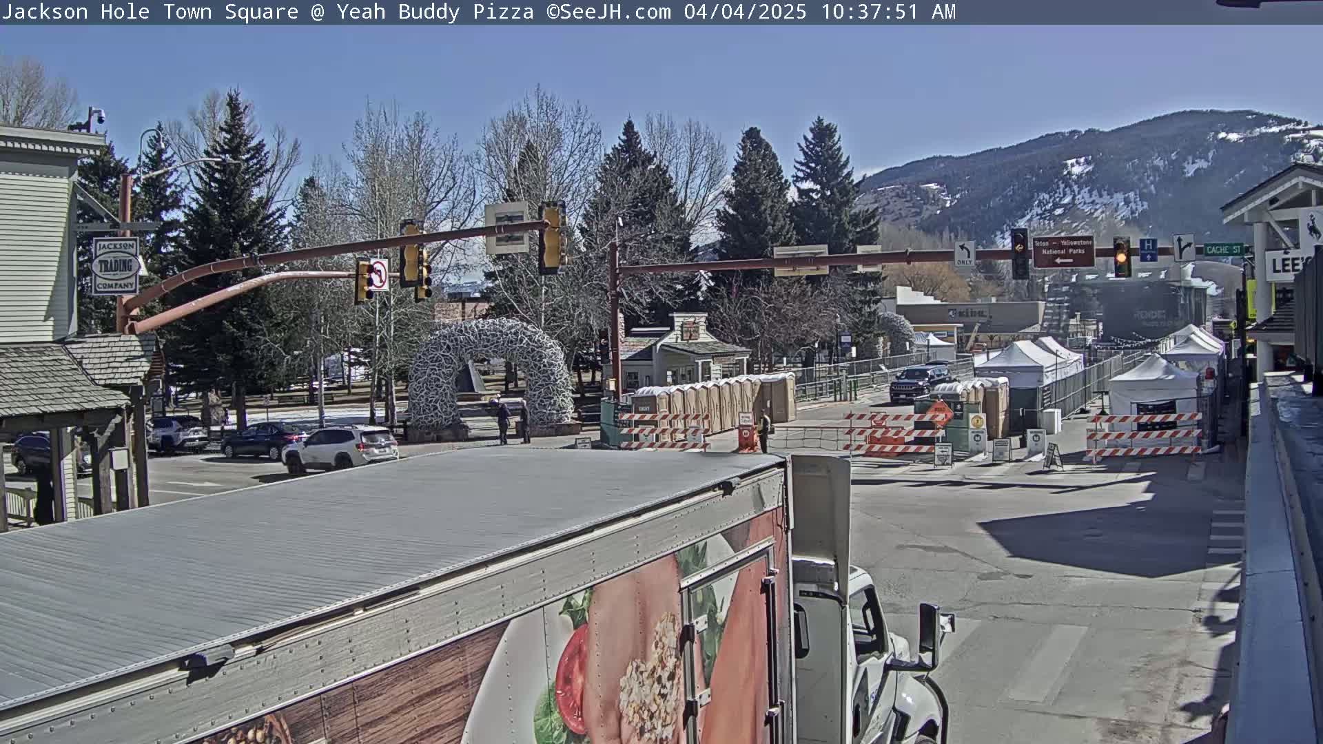 A sunny day in a town square shows several cars parked and driving, temporary structures, and a large truck in the foreground with a food advertisement on its side, all set against a backdrop of mountains.