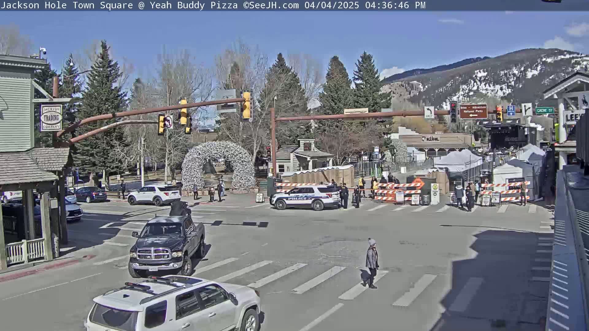 A sunny town square scene shows several vehicles, pedestrians, and police officers near barricades and tents under a clear blue sky, with snow-capped mountains in the background.