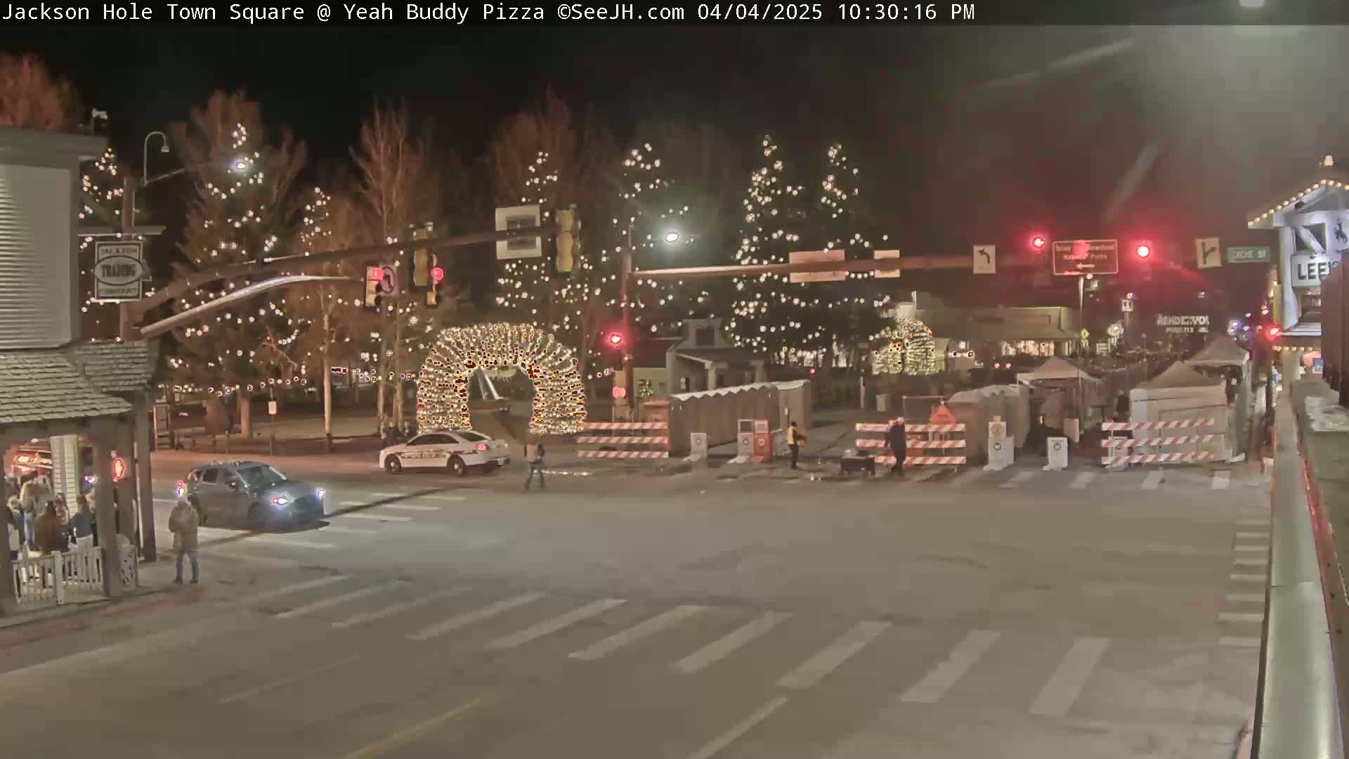 A nighttime view of a town square decorated with Christmas lights, featuring vehicles and pedestrians at an intersection.
