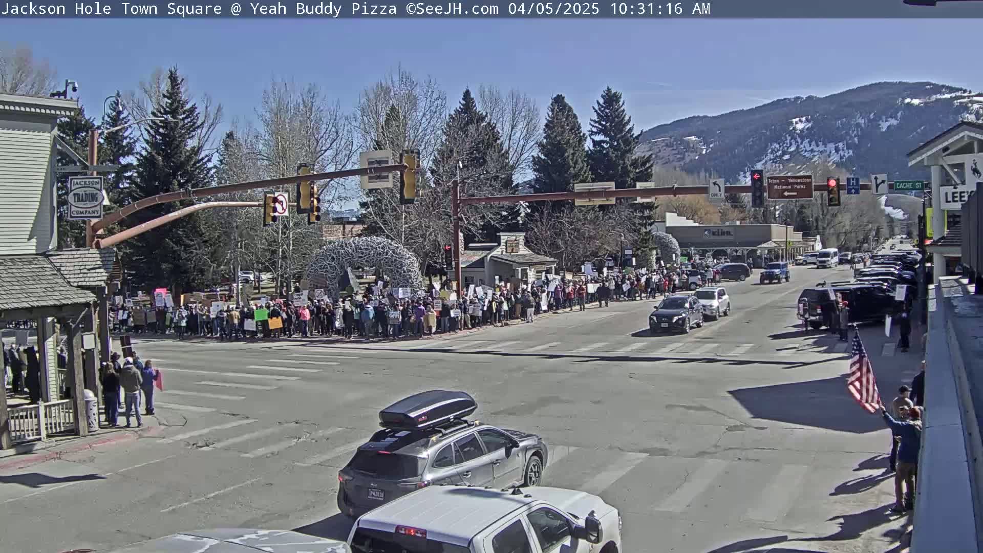 A large group of people are protesting in a town square on a sunny day, with cars driving on the surrounding streets and mountains visible in the background.