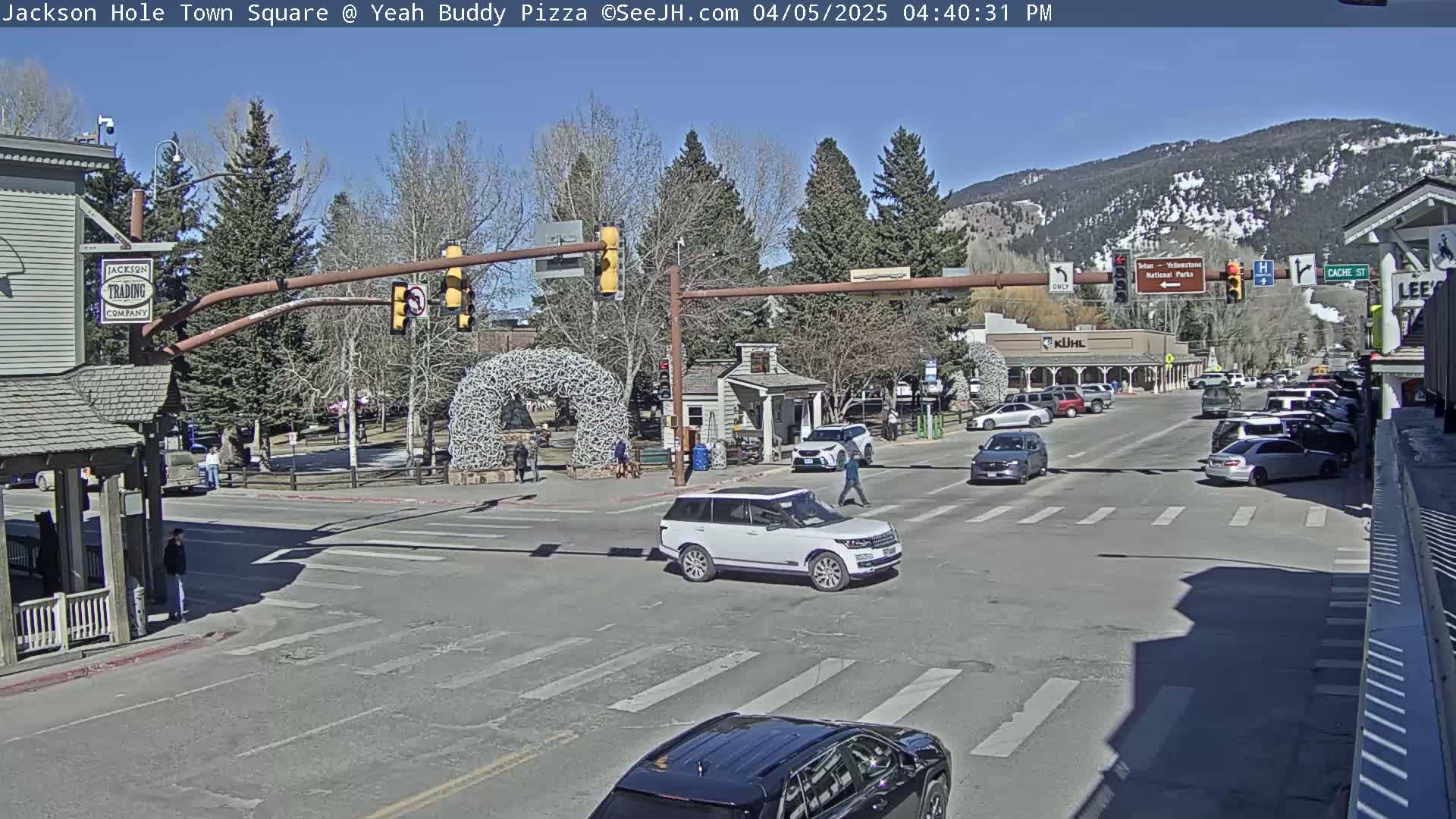 A sunny day shows a town square intersection with several cars and pedestrians, and a large, decorative arch in the background.