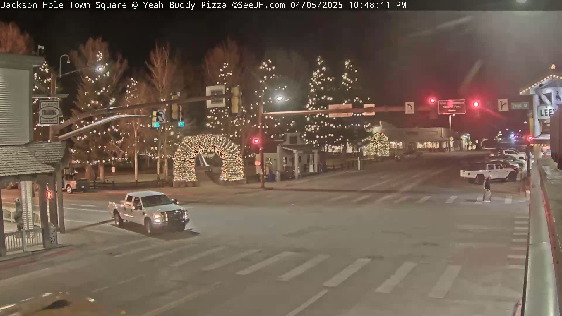 A nighttime scene of a town square with Christmas lights on trees and archways, a few vehicles, and pedestrians crossing a street.