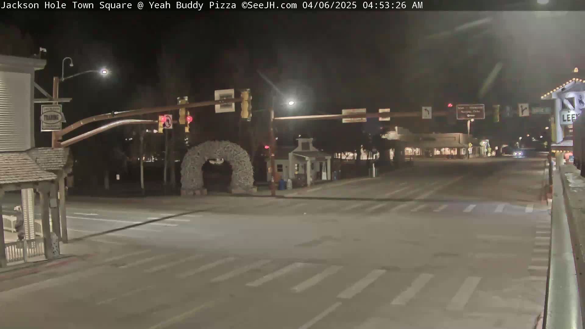 A nighttime, mostly empty town square intersection with crosswalks and traffic lights, featuring a decorative archway and some buildings.