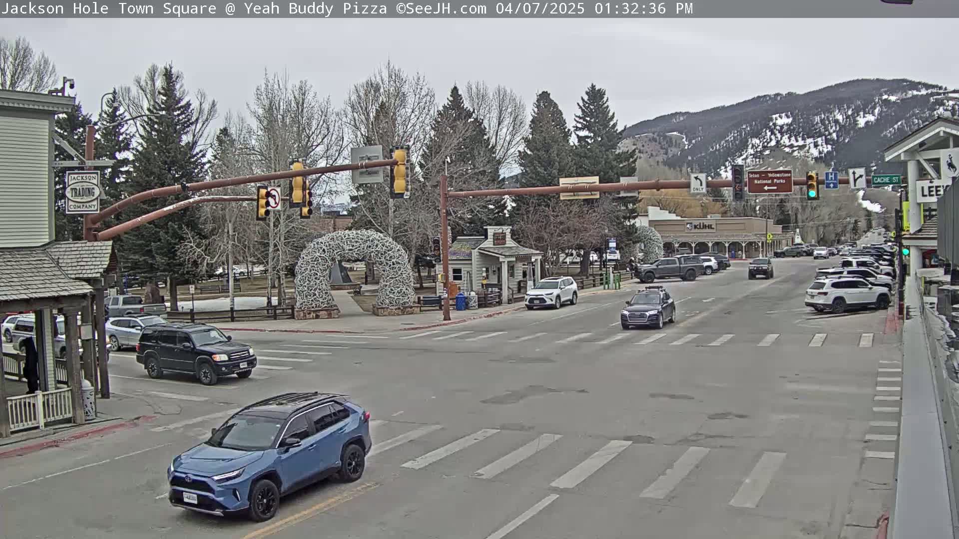 A town square scene on an overcast day shows a street intersection with several cars, buildings, and trees, with mountains in the background.