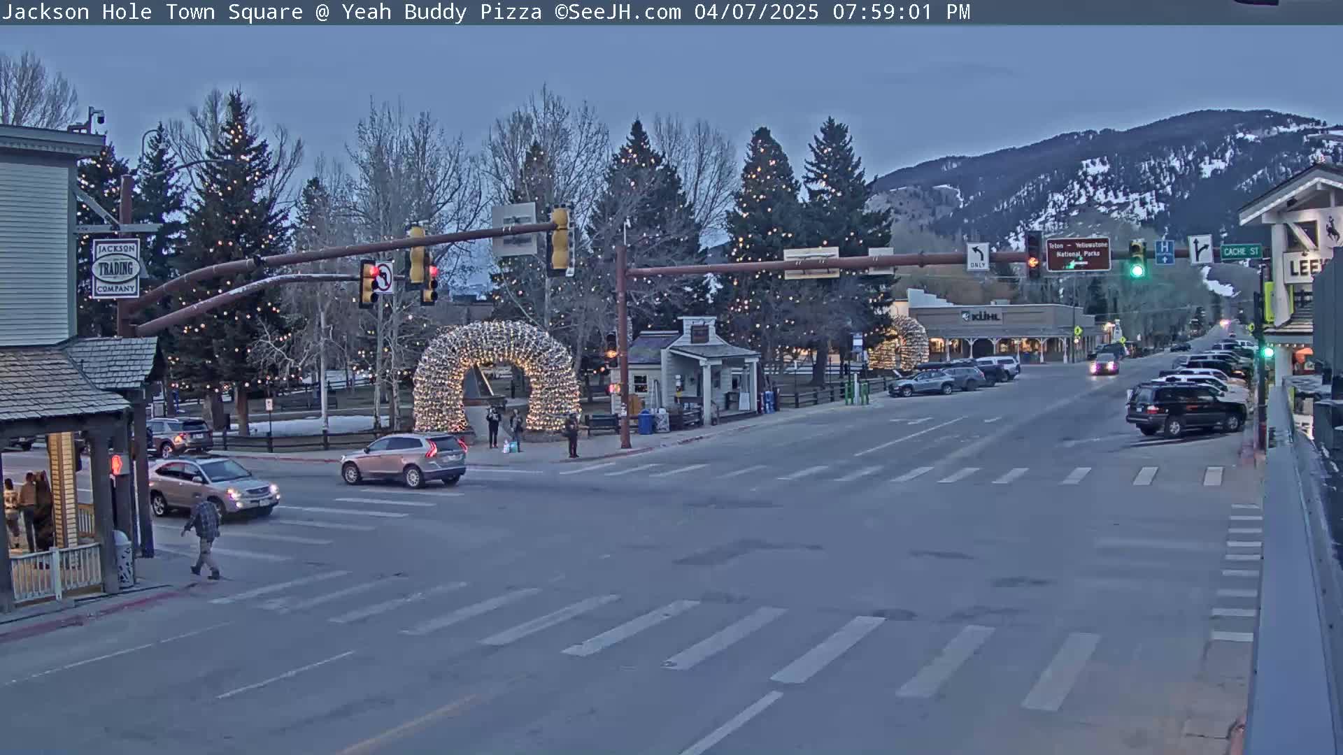 A town square at dusk, illuminated by string lights on trees and arched structures, shows several cars at a crosswalk and pedestrians on the sidewalk under a twilight sky with mountains in the background.
