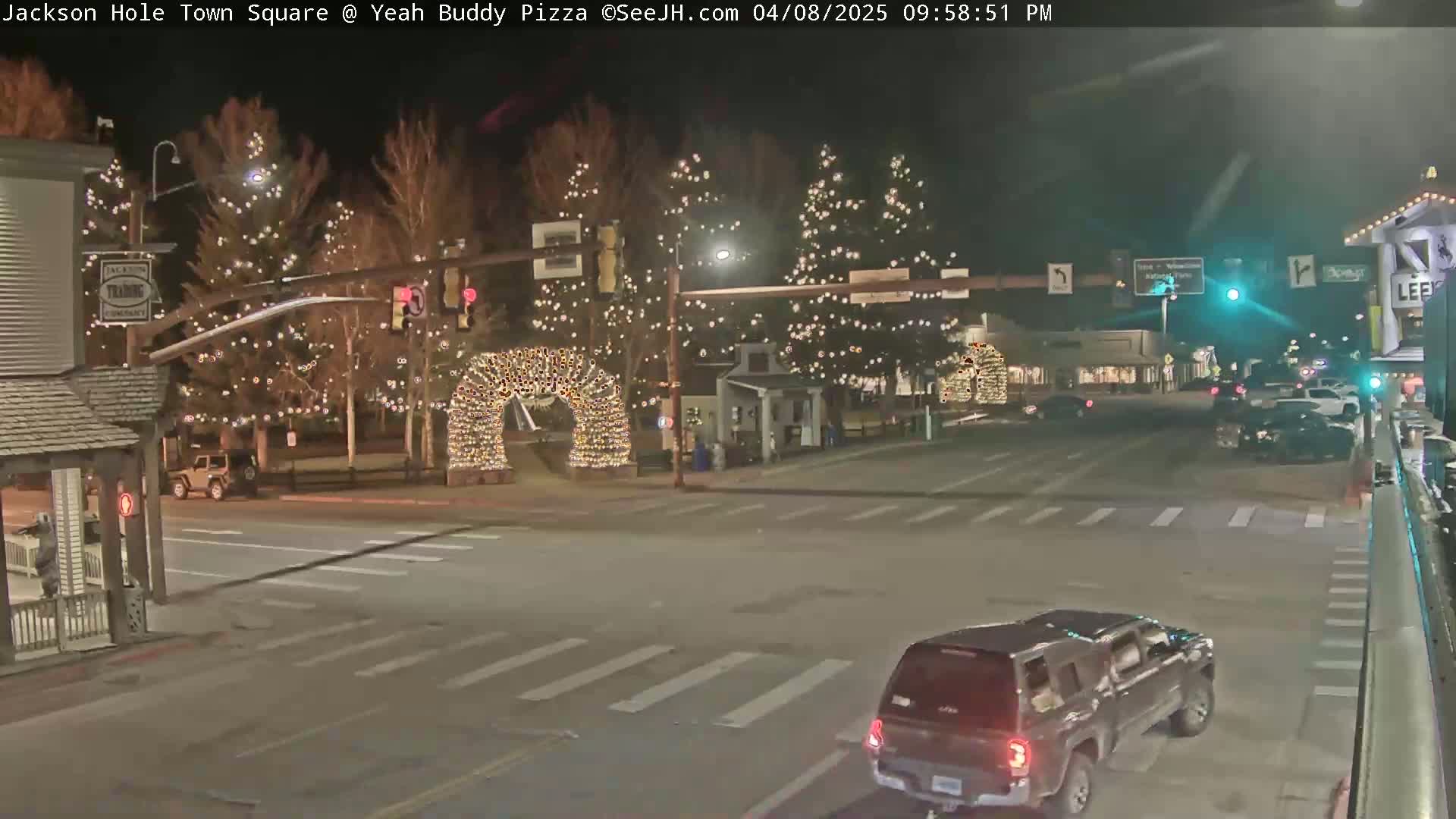 A nighttime view of a town square with Christmas lights strung on trees and an archway, a few cars driving on the road, and buildings lining the street.