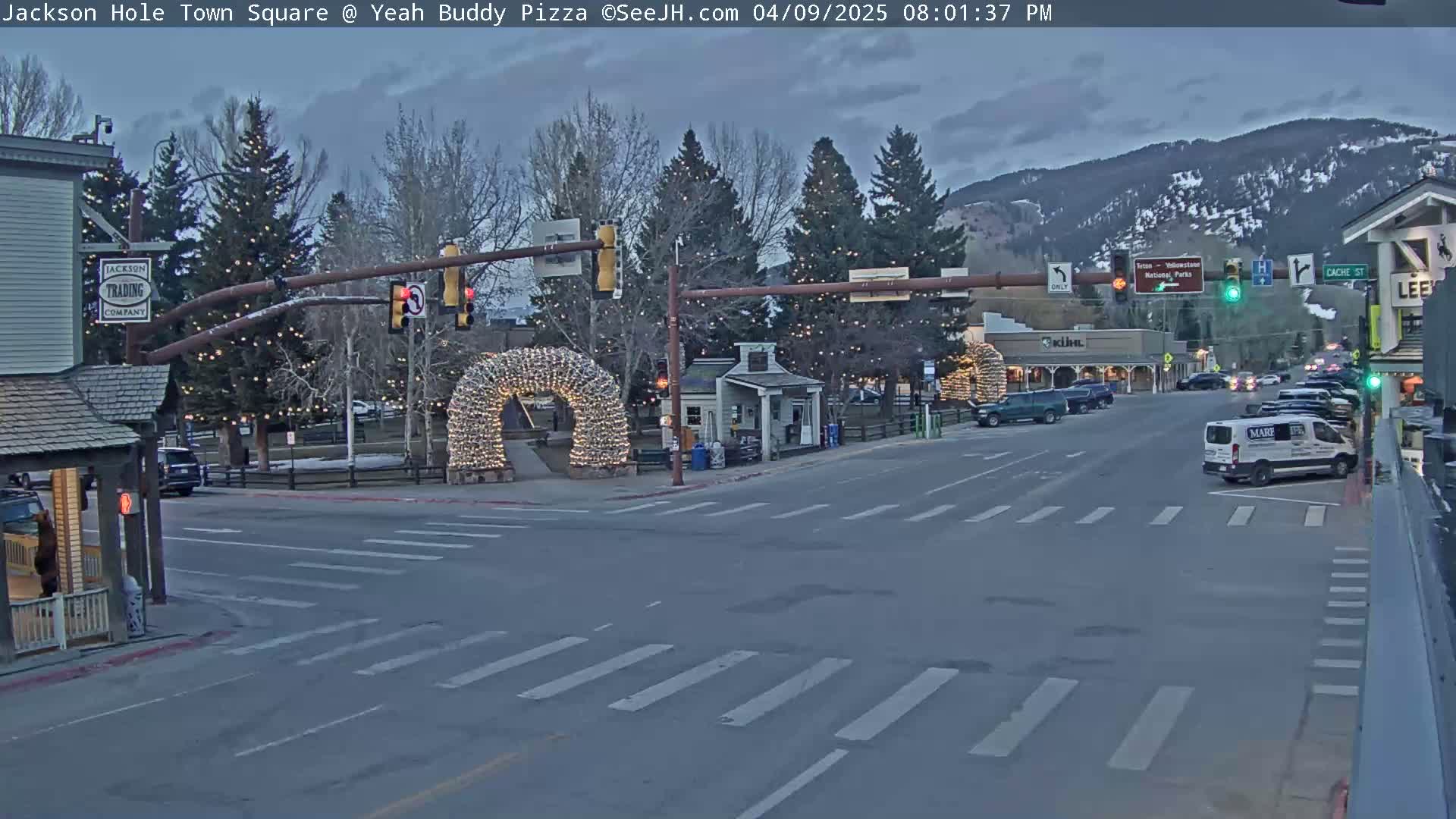 A town square at dusk, with a few cars and a light-up archway, under an overcast sky.