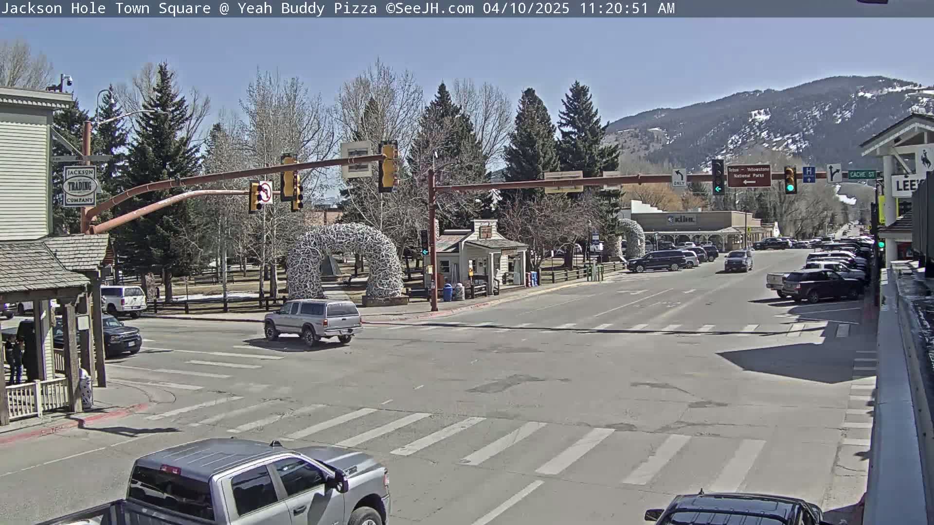 A sunny town square scene shows a street intersection with cars, buildings, and a large antler arch, with mountains in the background.