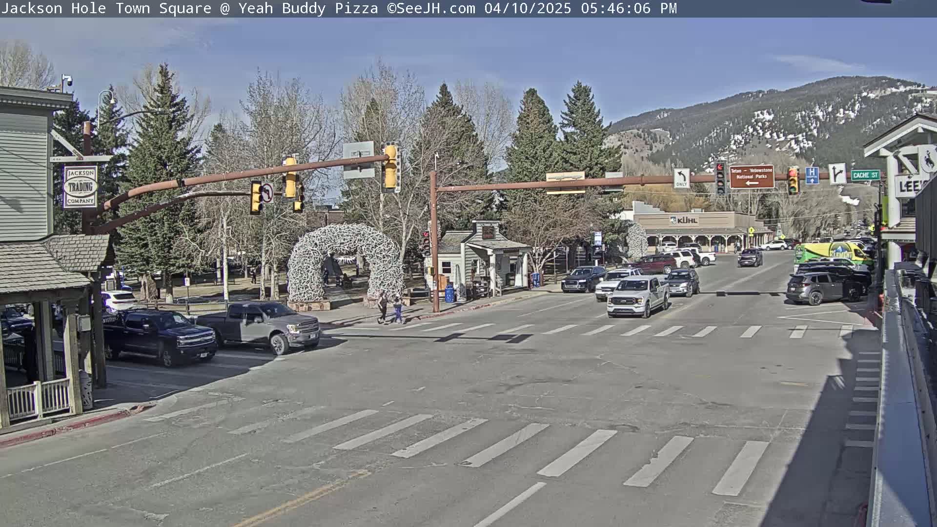 A sunny town square scene shows a street intersection with several cars and pedestrians,  backed by buildings and mountains.