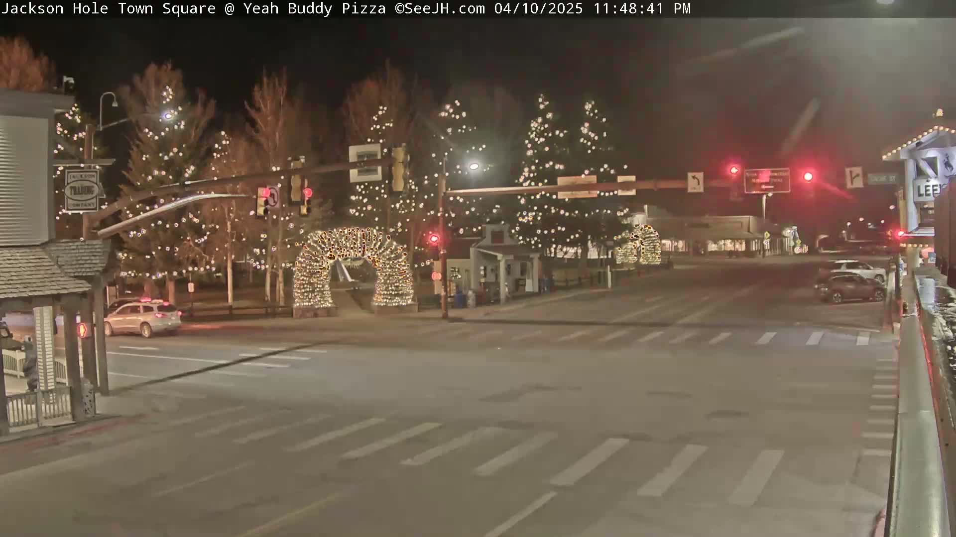 A nighttime street scene shows a town square with Christmas lights strung on trees, an arched light display, and a few cars at an intersection.