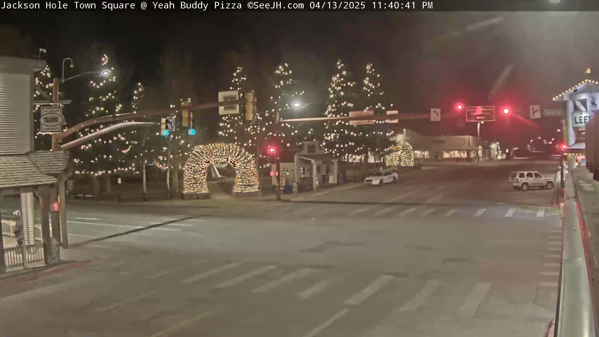 A nighttime view of a town square with Christmas lights on trees and an archway, a few cars, and traffic lights.