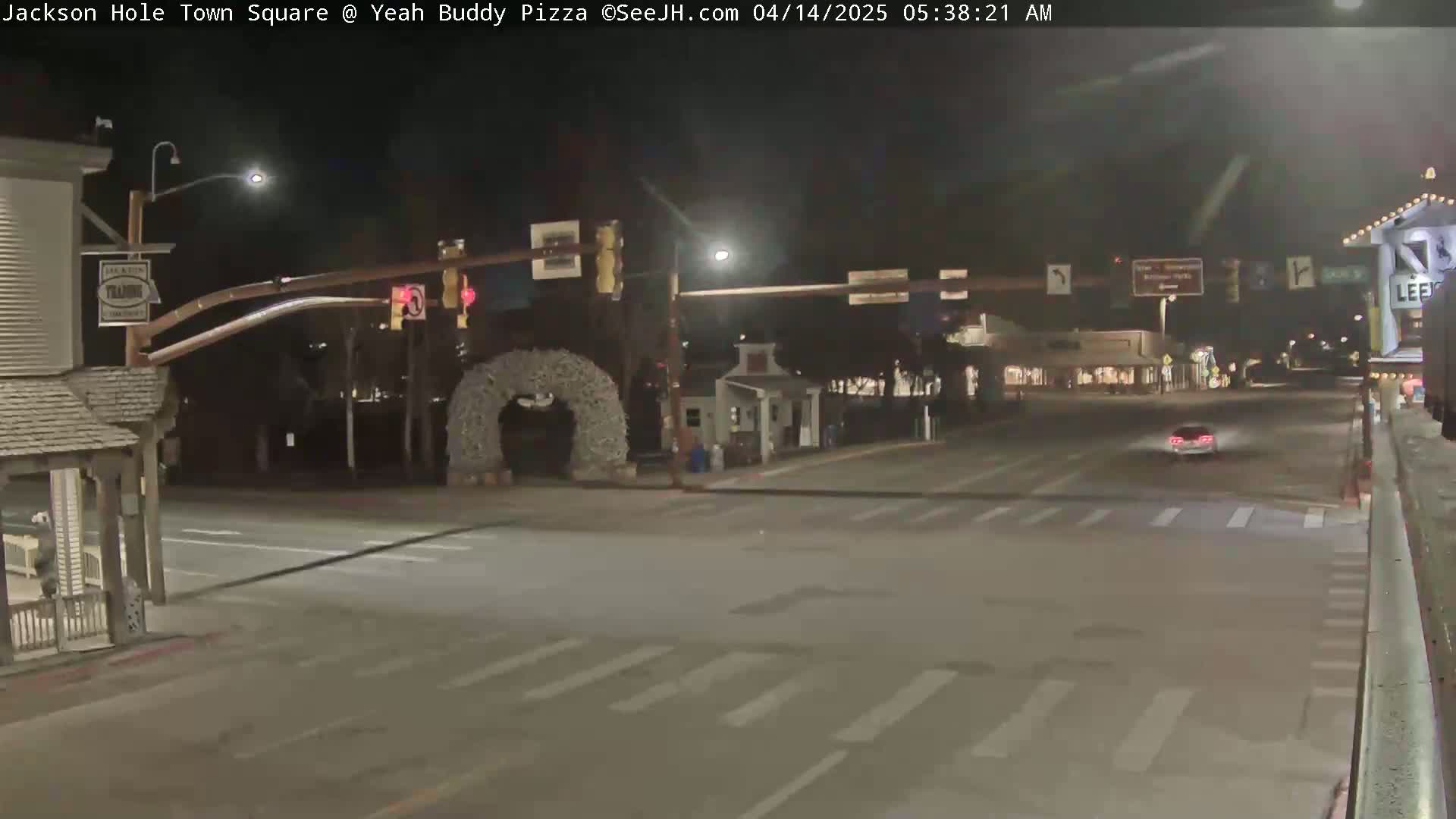 A nighttime view of a mostly empty town square intersection with traffic signals and a single vehicle driving through.