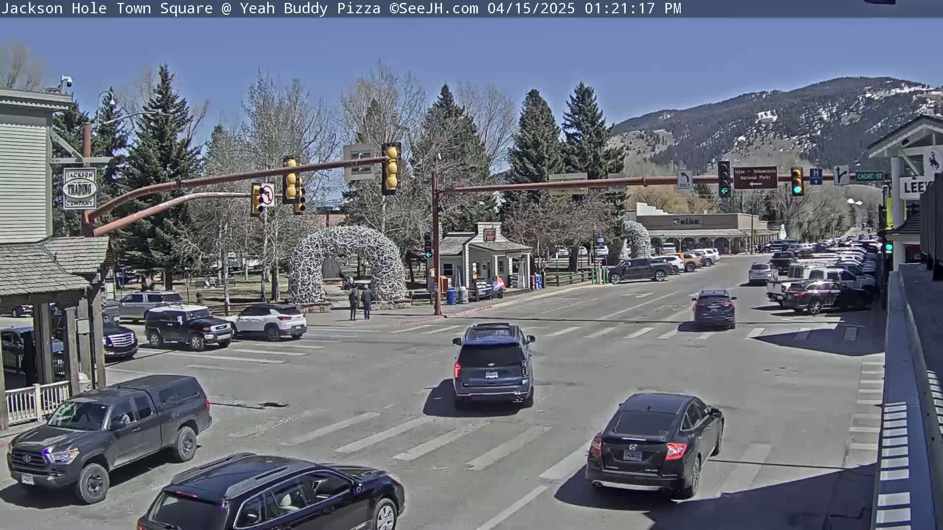A sunny town square with several cars at a traffic intersection, surrounded by buildings and trees, with mountains in the background.
