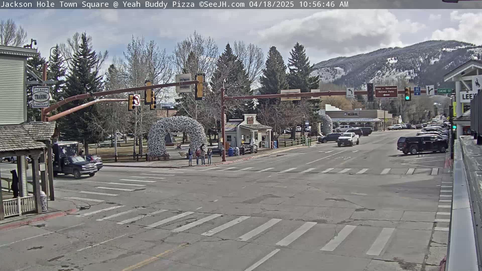 A mostly sunny town square scene shows a few cars at an intersection, pedestrians near a large metal arch sculpture, and buildings with trees in the background under a partly cloudy sky.