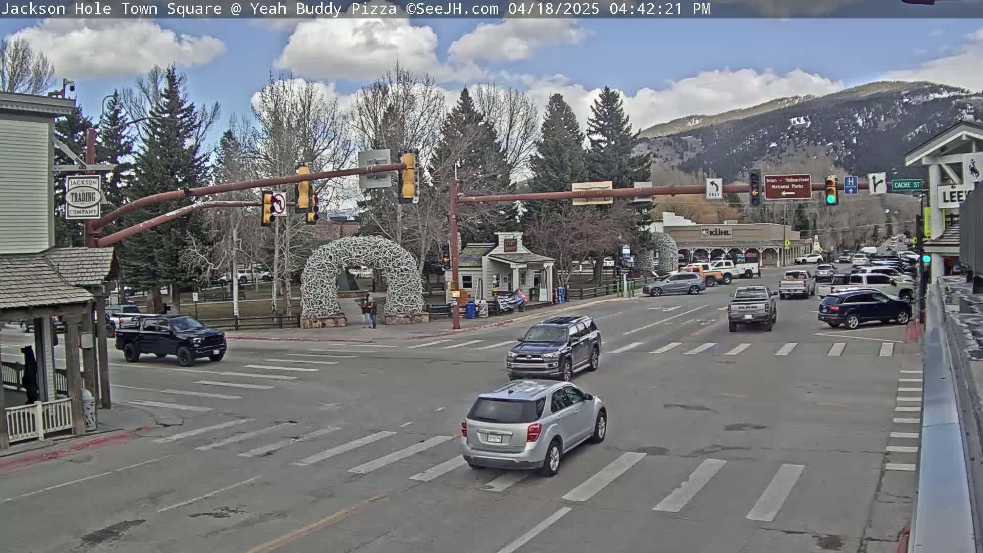 A partly cloudy day in a town square with several vehicles at a traffic intersection, surrounded by buildings and trees, with mountains visible in the background.