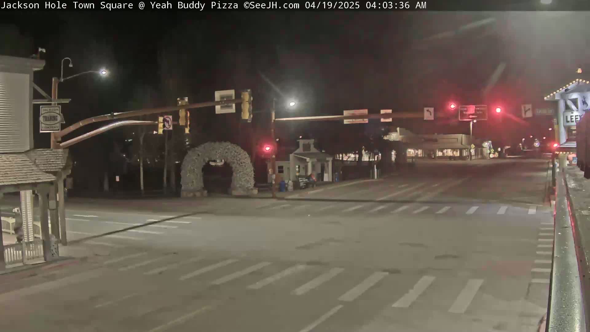 A nighttime view of a mostly empty town square intersection with traffic lights, some buildings, and a decorative arch.