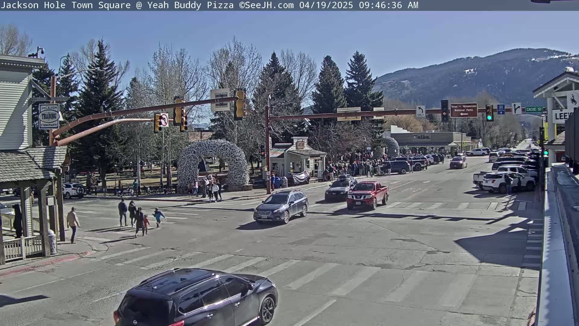 A sunny town square with pedestrians crossing a street, several cars stopped at intersections, and mountains in the background.