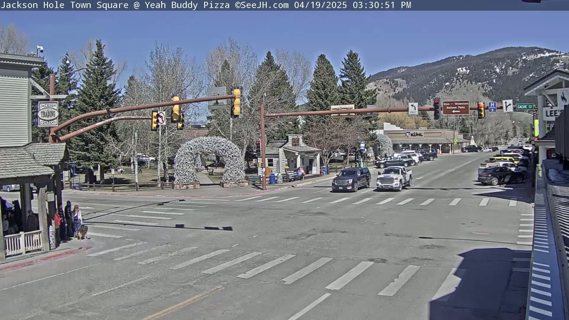 A town square with a few cars and pedestrians, under a clear blue sky,  backed by snow-capped mountains in the distance.