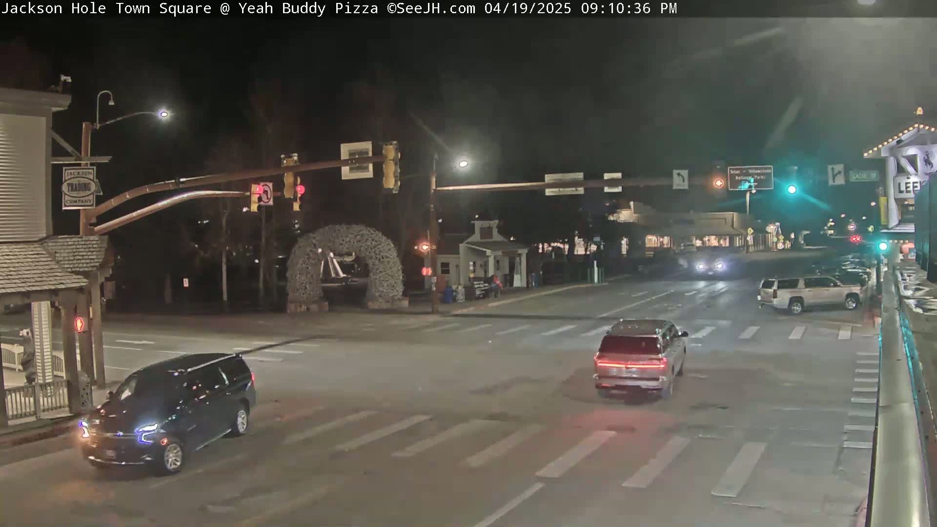 At night, a dark-colored SUV and a light-colored SUV are driving through a town square intersection under traffic signals.