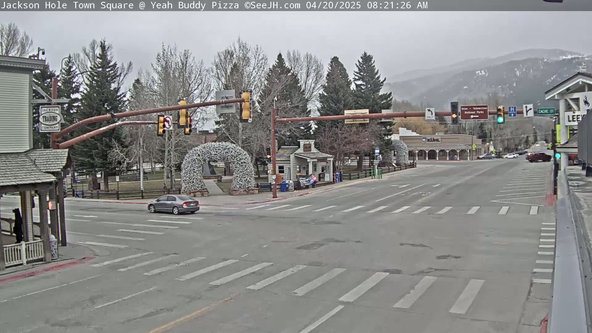 A mostly cloudy day shows a town square intersection with a few cars, bare trees, and a large antler arch.