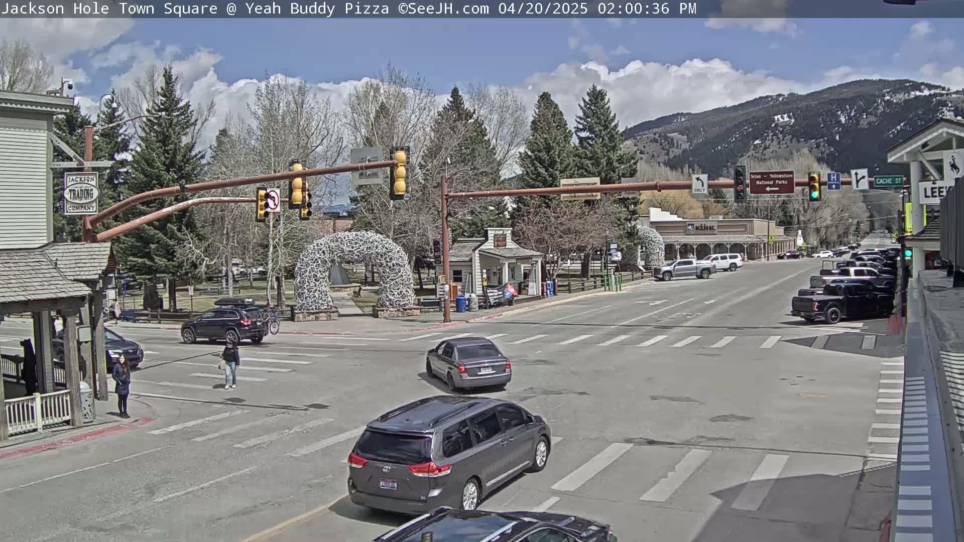 A partly cloudy day shows a town square intersection with several vehicles and pedestrians, mountains in the background.