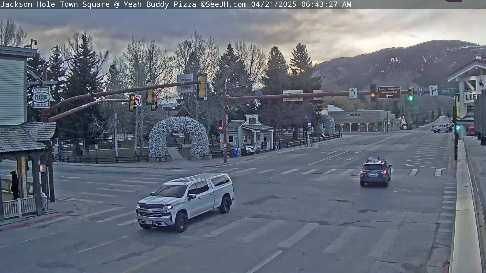 A mostly cloudy morning scene shows a town square intersection with several vehicles, including a silver pickup truck,  and trees, with a mountain range visible in the background.