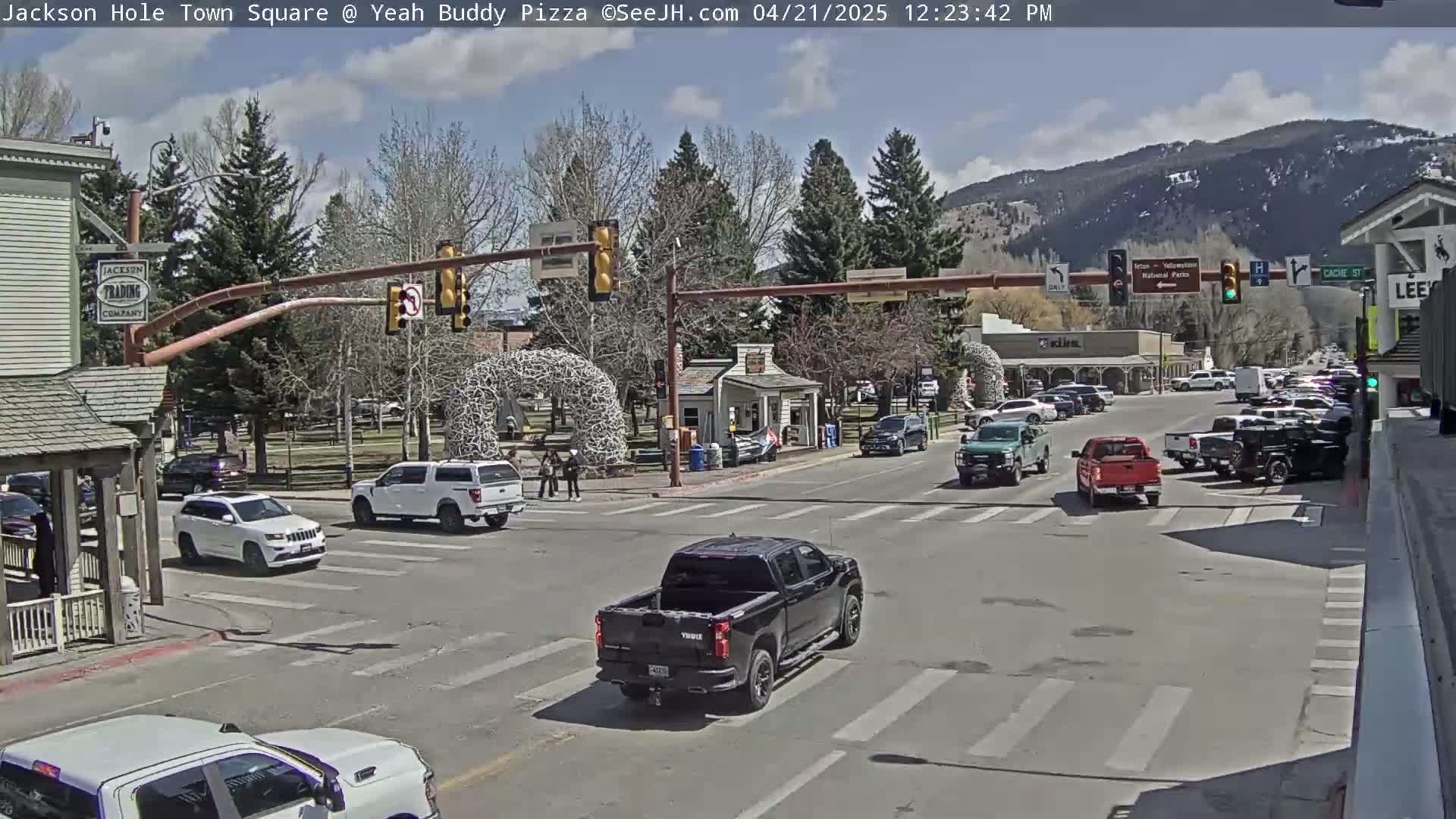 A partly cloudy day in a town square shows several vehicles at an intersection, with mountains in the background.