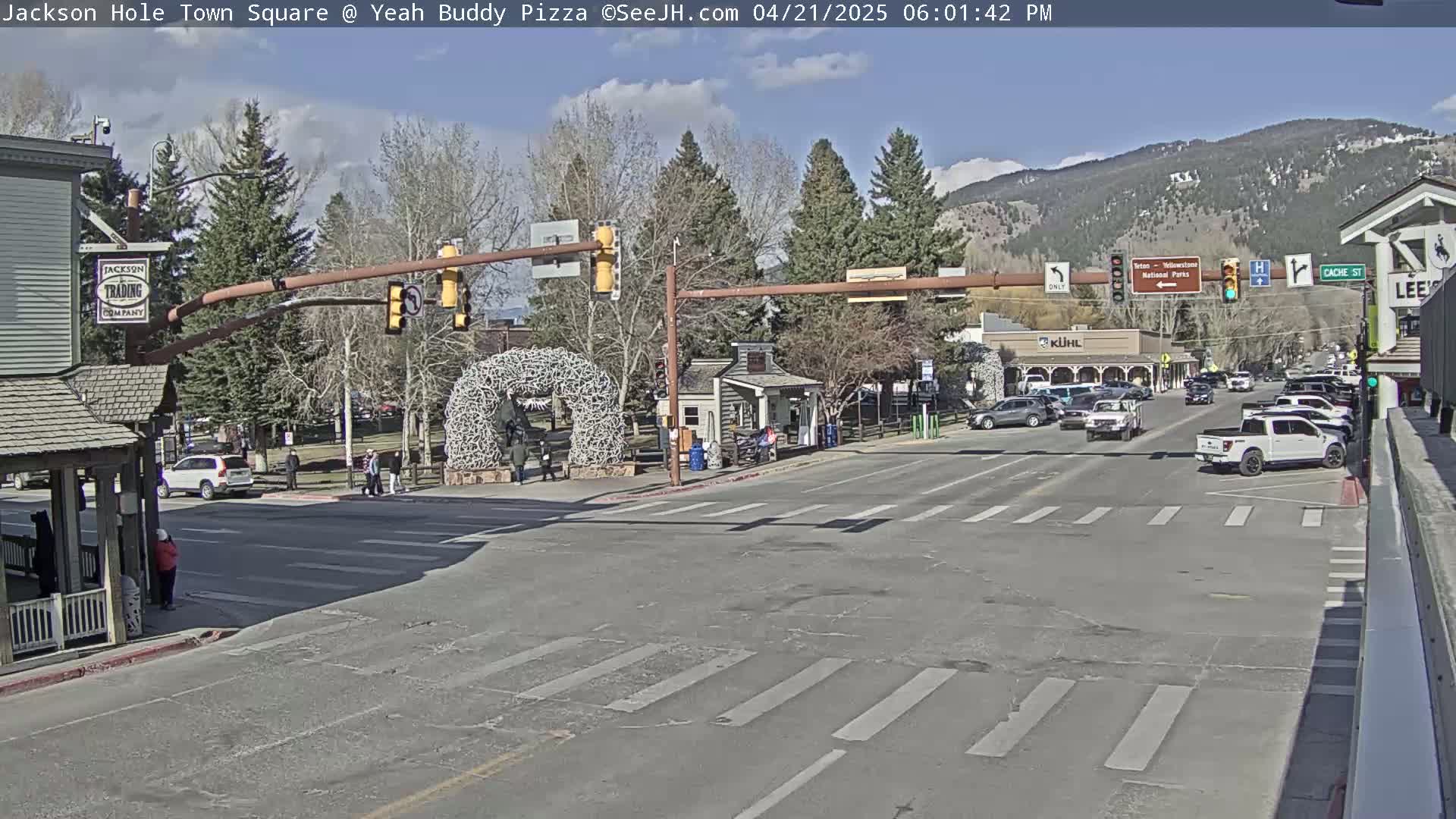 A sunny town square intersection with cars and pedestrians, under a mostly clear sky, backed by snow-capped mountains.