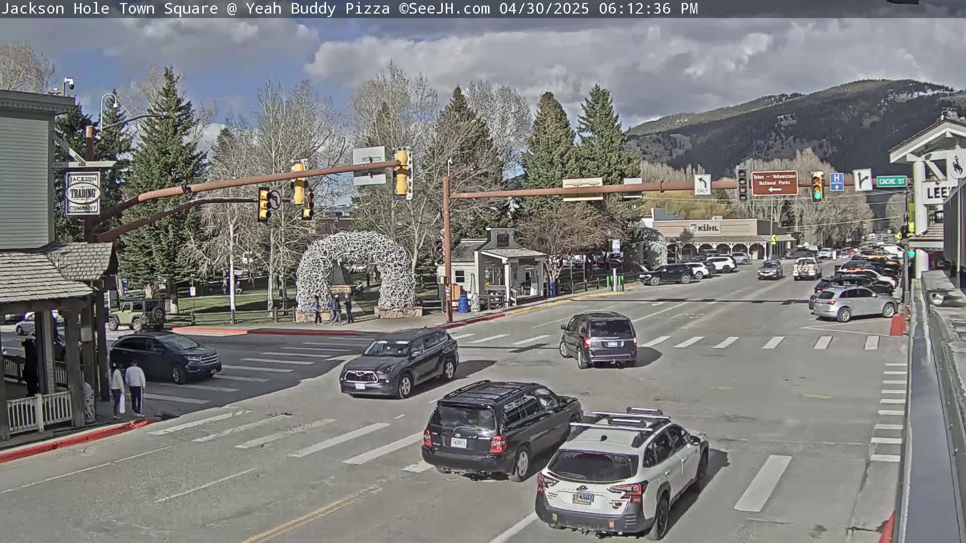 A sunny town square intersection with several vehicles and pedestrians, under a partly cloudy sky, is bordered by buildings and trees, with mountains in the background.
