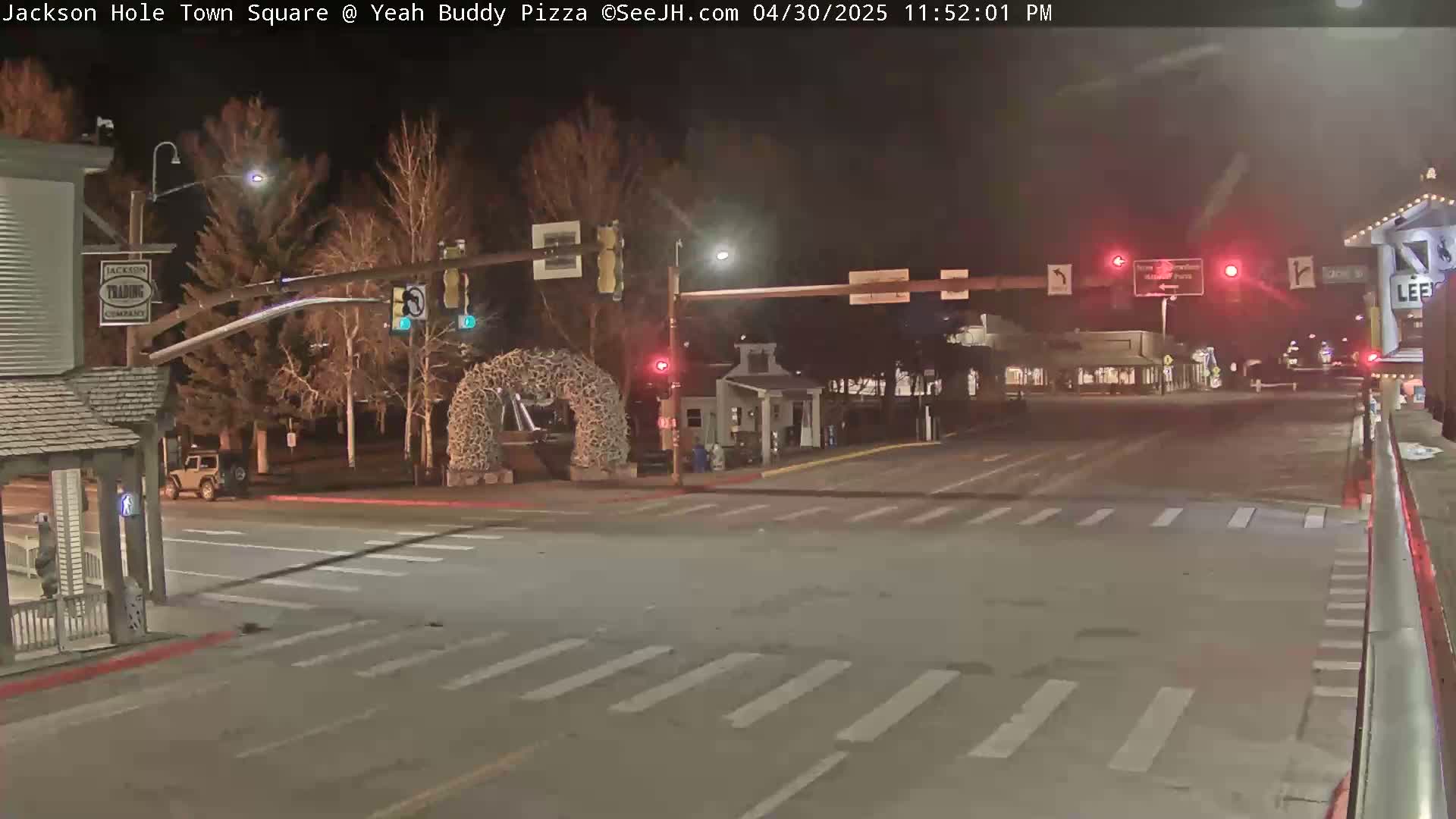 A nighttime street scene shows a sparsely populated town square with a vehicle at an intersection under traffic signals, and a large antler arch in the background.