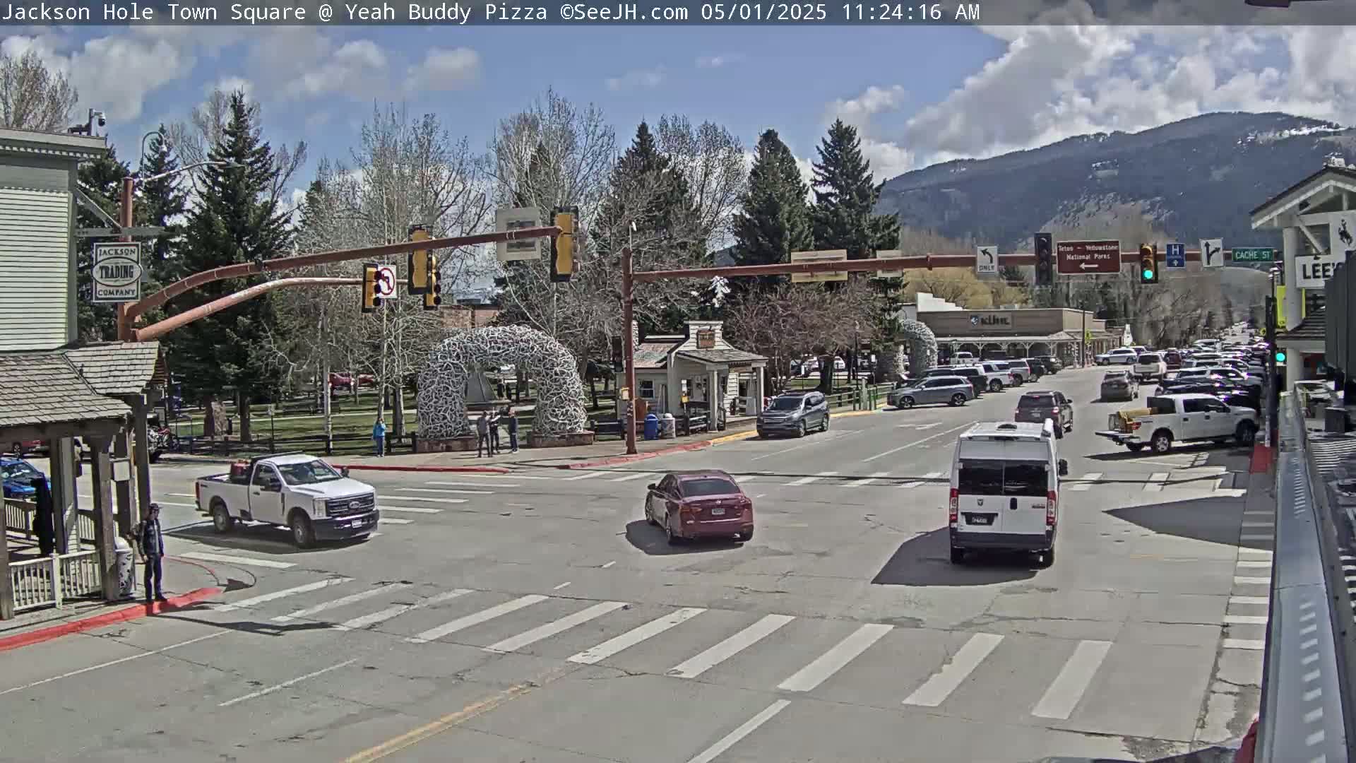 A partly cloudy day shows a town square with cars at a four-way intersection, several parked cars, and trees lining the streets under a partly cloudy sky with mountains in the background.