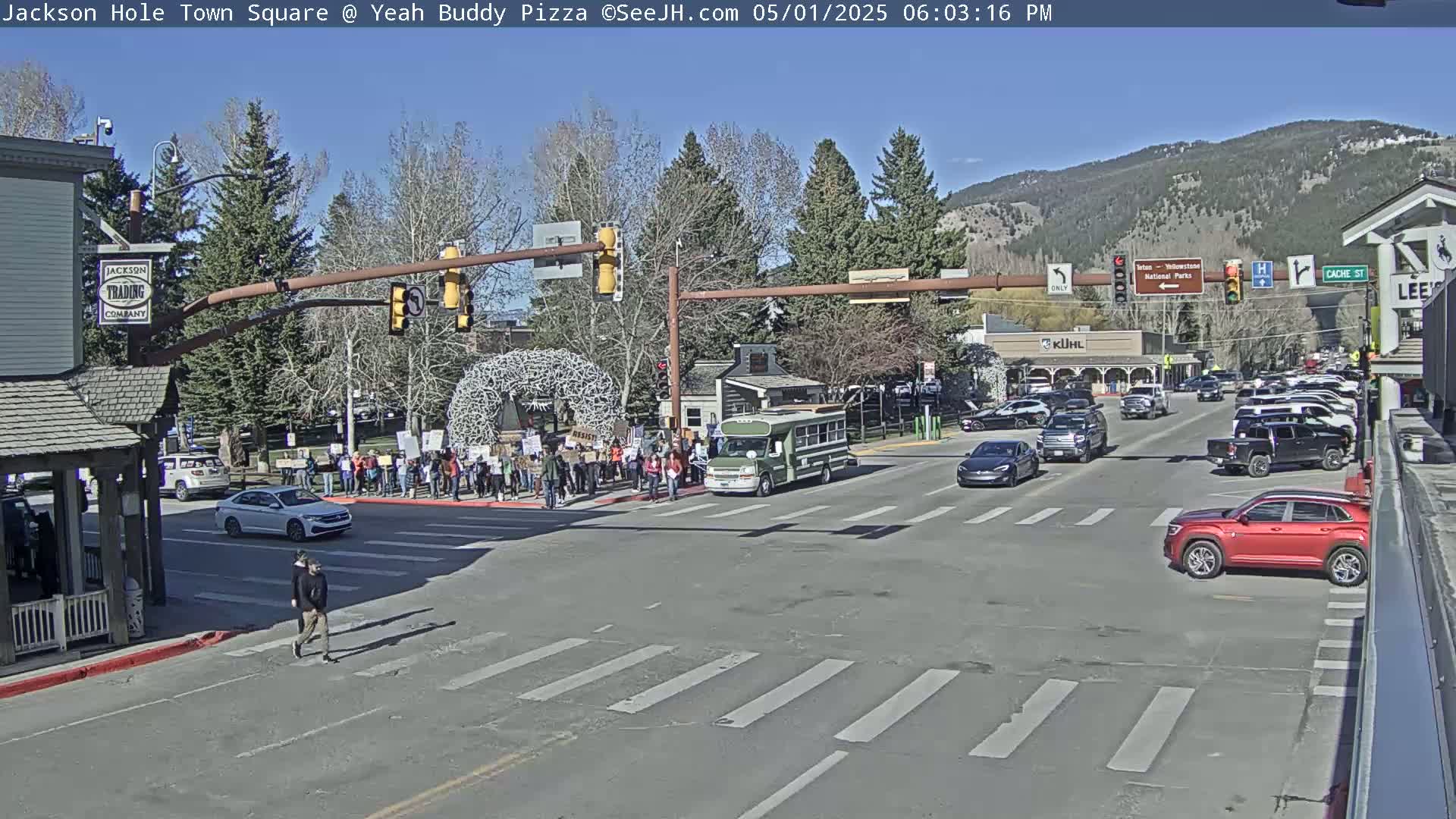 A sunny day in a town square shows a crowd of people standing at a crosswalk, several cars driving through an intersection, and a green van stopped at the traffic light.