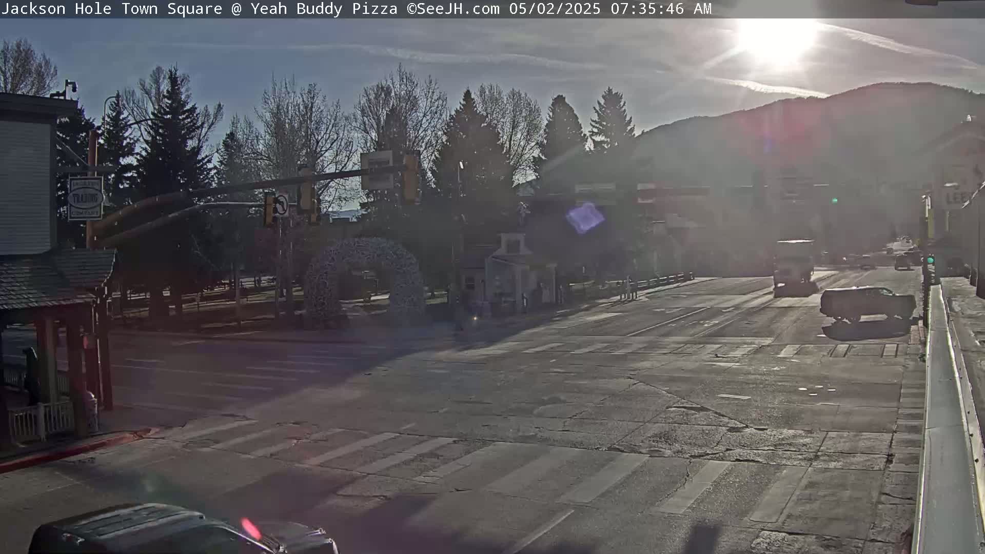 A sunny morning scene shows a town square intersection with several vehicles, pedestrians, and trees, with mountains in the background.