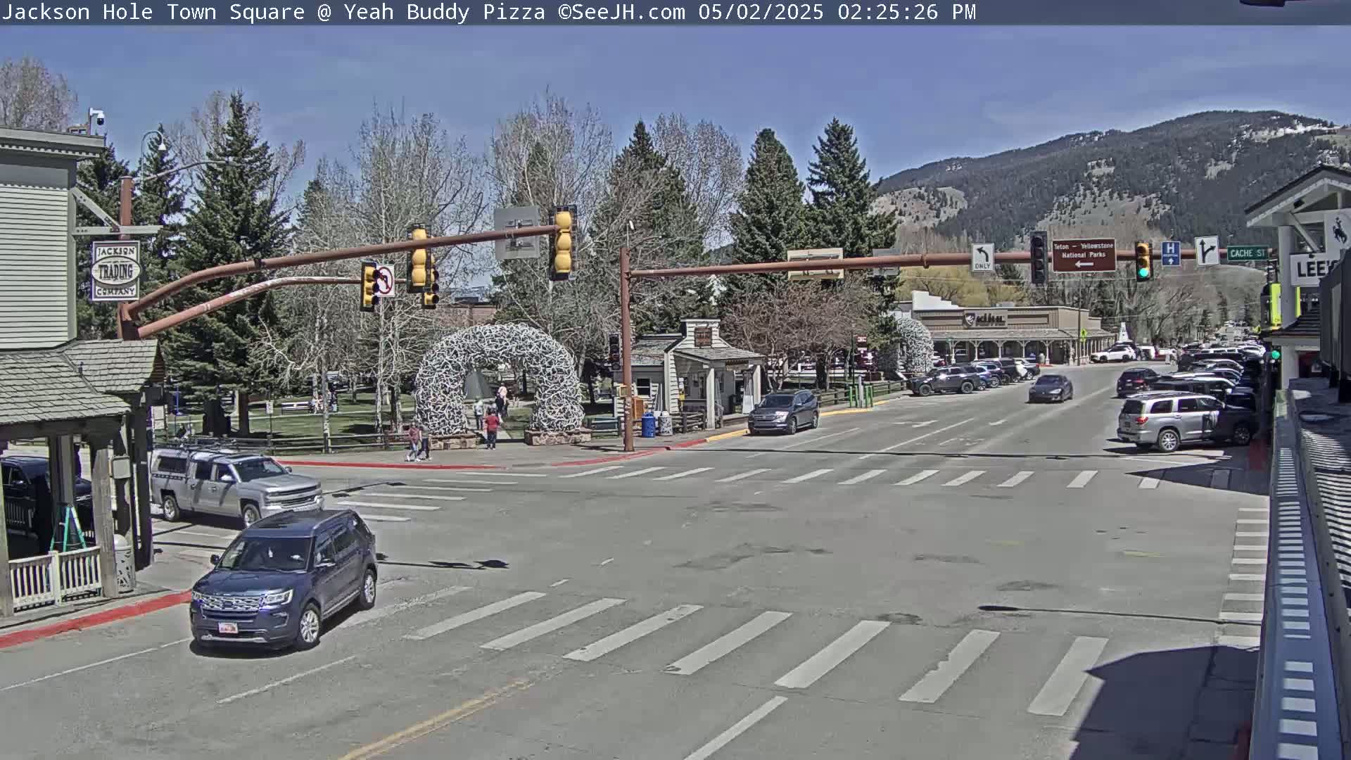A sunny town square with cars parked along the side of the road and a few cars driving through the intersection, under a clear sky and mountains in the background.