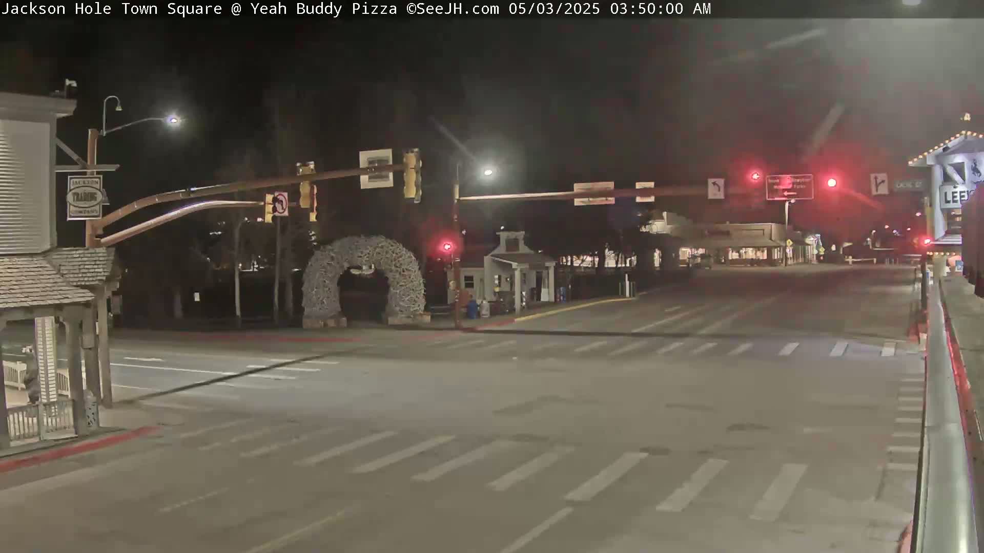 A nighttime view of a mostly empty town square intersection with traffic lights, street markings, and a decorative archway.