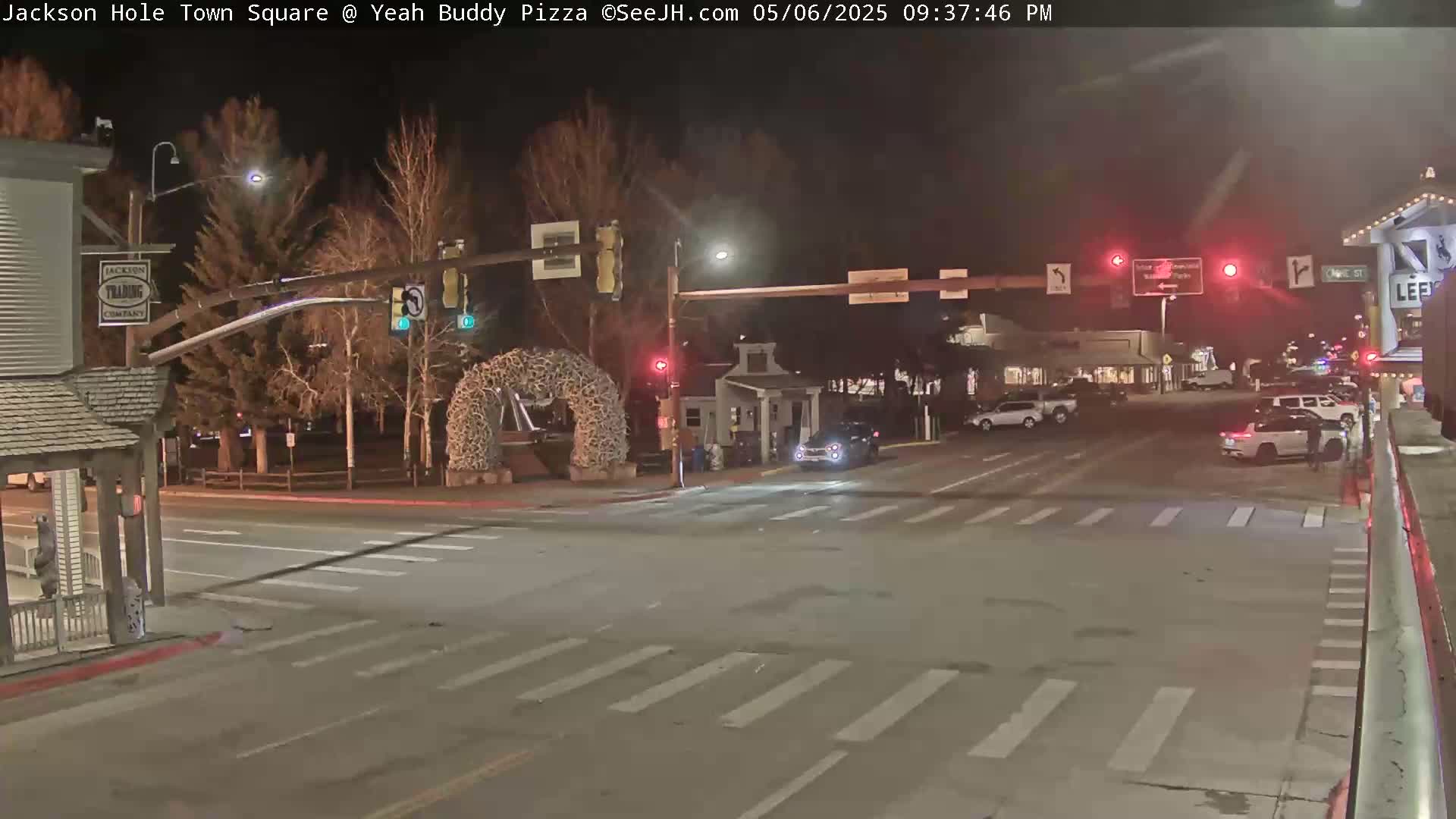 A nighttime street intersection, with sparse traffic and bare trees under dark skies, shows a crosswalk and traffic lights.